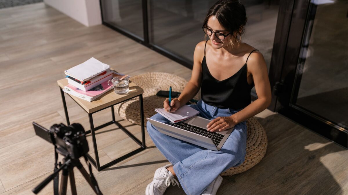 Young woman studying indoors with a laptop and camera setup, symbolizing remote learning.