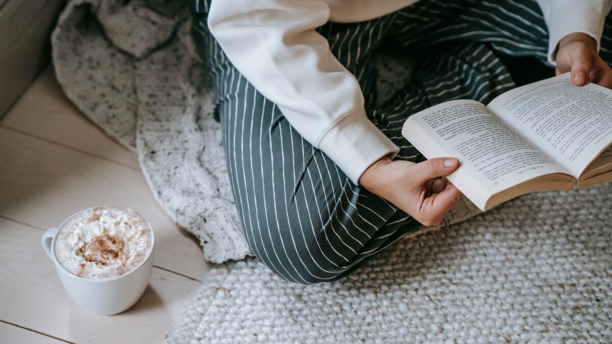High angle of crop anonymous woman sitting on soft blanket on floor and reading literature with mug of coffee