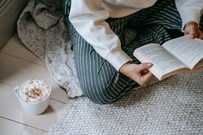 High angle of crop anonymous woman sitting on soft blanket on floor and reading literature with mug of coffee