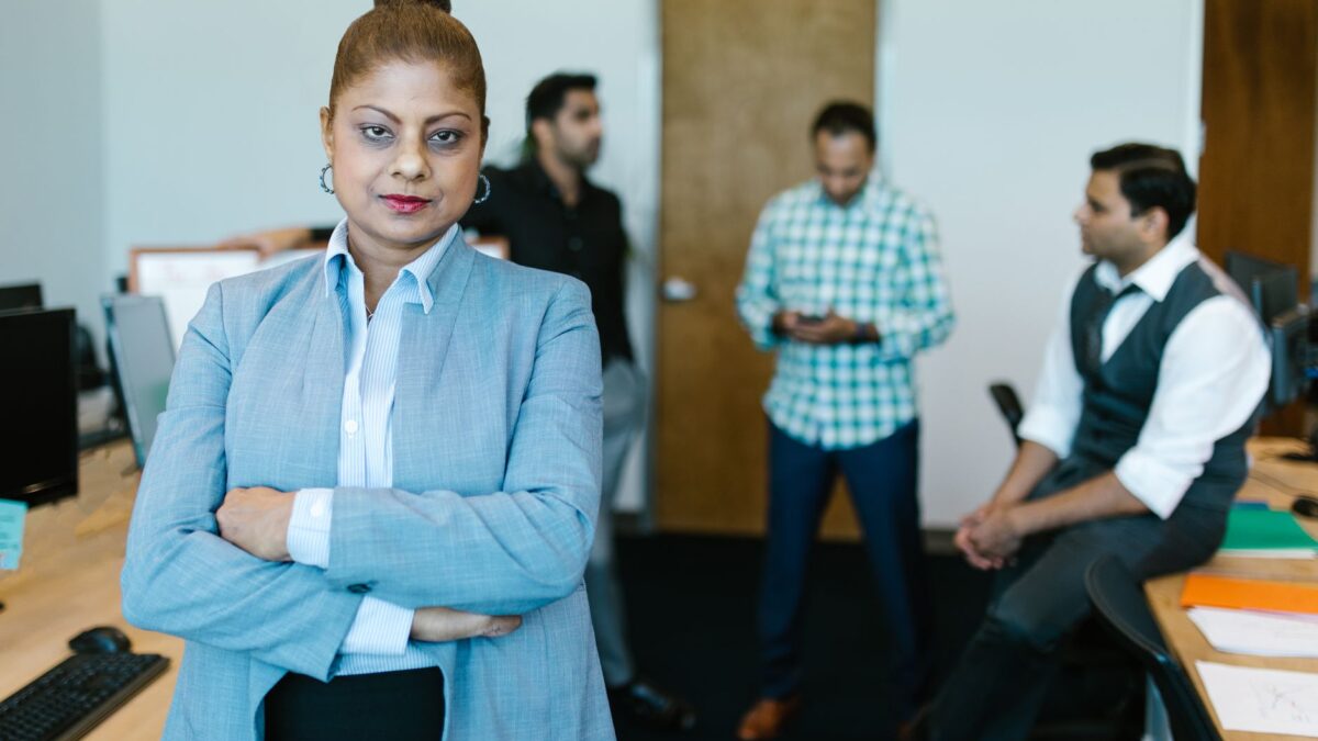 Confident businesswoman standing in modern office with colleagues in the background.