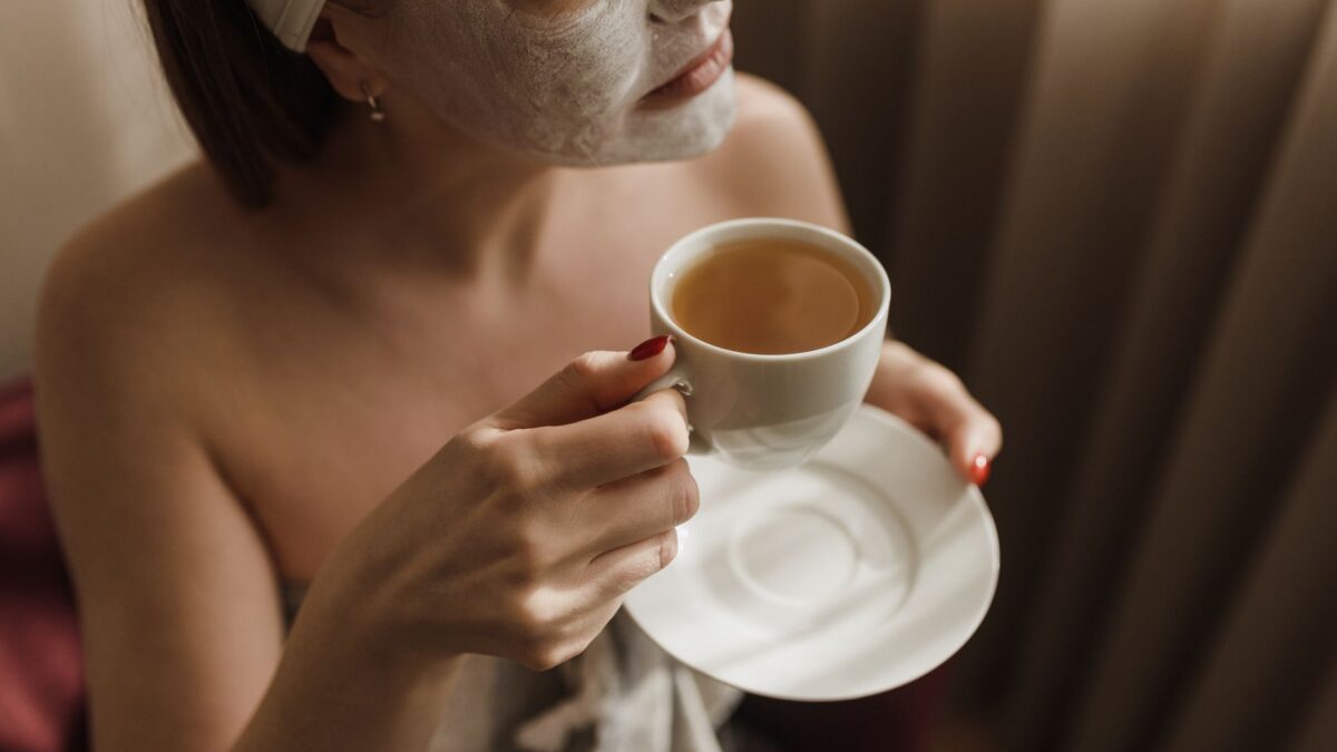 A woman with a facial mask enjoys a warm beverage during a skincare routine.