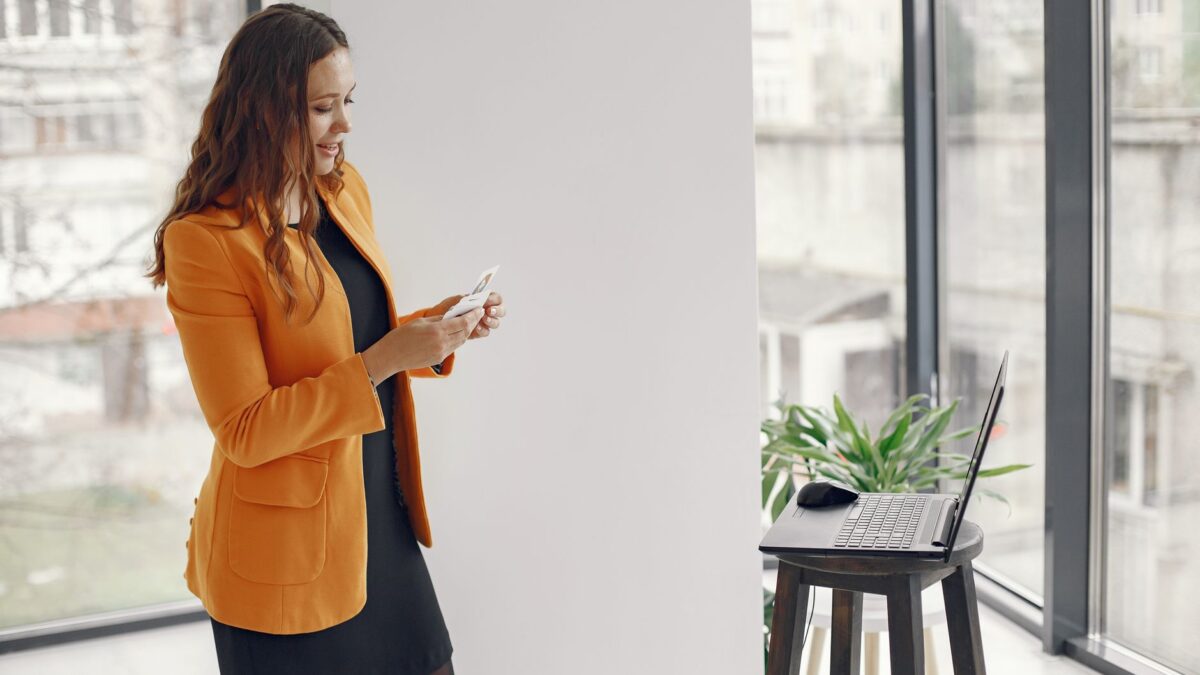 Businesswoman with orange jacket holds cards and stands by laptop in modern office.