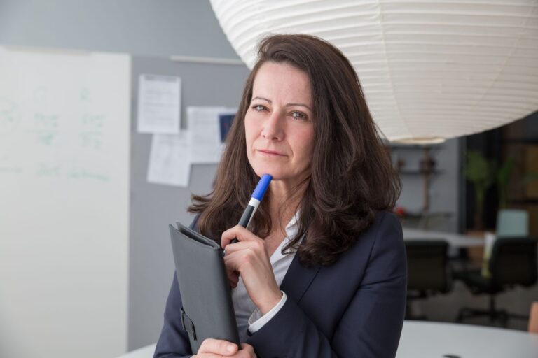 Businesswoman in corporate attire posing with a pen in a modern office setting.