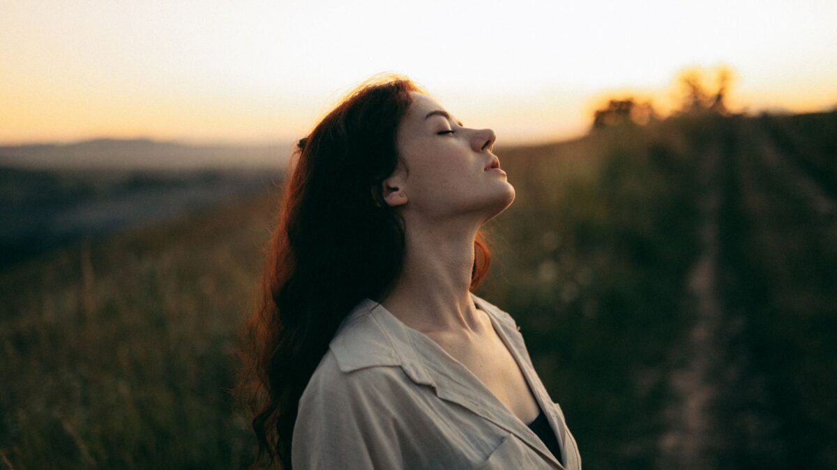 Woman standing with eyes closed in a field during sunset, exuding tranquility and peace.