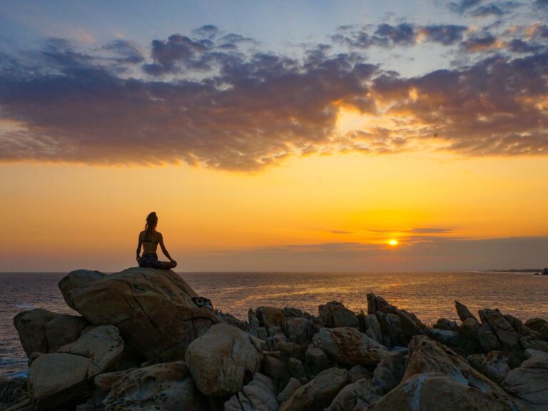 A woman meditates on a rocky shore during a beautiful ocean sunset, creating a peaceful scene.
